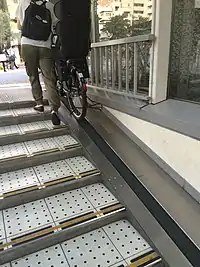 Shallow steps with a sliding black-rubber motorized ramp alongside; a cyclist pushing a bike to the top of the ramp