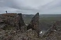 A hiker stands on a mountain summit near a rocky outcropping, with a vast landscape with lakes in the background