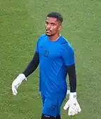 Photograph of a man in a blue jersey and shorts with a black long-sleeve beneath it. He is wearing white goalie gloves.
