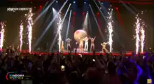 Six people, one in a nude suit and five with shirts pulled over their heads, jumping around on a stage with a giant breast globe behind them.