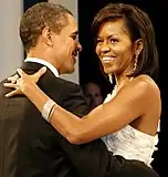 Barack and Michelle Obama are dancing arm-in-arm and smiling. She is wearing a white dress, a large ring, long earrings, and a bracelet. He is wearing a black tuxedo.
