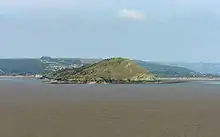 Green covered rocky land in expanse of sea. Hills behind.