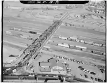 An aerial view of a rail yard, in which a number of freight train cars are visible, predominantly boxcars. A road bridge crosses over the rail yard and connects to a highway in the background.