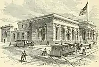 An Egyptian-Revival-style building on a street corner, with doric columns on its façade and a large flag flying atop a mast. Pedestrians are seen navigating two horse-drawn trams, which travel on lines that bisect intersecting streets.