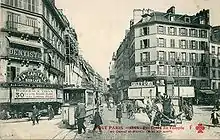 Sepia picture postcard of a commercial street scene, taken from a crossroads, looking down the street. A two-car train is in the foreground, head-on. Several pedestrians are crossing towards it.