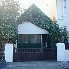 An extremely small workshop adjoining a house and standing behind a wooden gate with white gateposts. It has green double doors with an open door to their right. Above this is a white-painted wall which is heavily draped with ivy, although a circular louvre is clearly visible through this. The roof is steep and pointed.