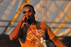 Gucci Mane holds a microphone onstage. He is wearing his ice cream cone pendant necklace but does not yet have the tattoo on his face.