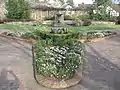 Photograph of a formal garden at the Luther Burbank House and Gardens, featuring brick walkways and planterboxes, a small lawn, a fountain and pool, low stone walls, and wooden trellises.