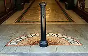 Close-up photograph of a black Victorian-style bollard in the centre of the arcade walkway. A semi-circular mosaic on the floor spells out the letters "Queen's Arcade" around the bollard.