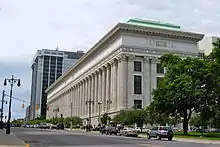 A tall white stone building with a colonnaded facade and intricate decorations on the stonework, much longer along the street to its left then the side facing the camera. There are trees in front of it on the right and a taller, more modern building behind it.