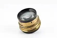 A small brass camera lens sits at an angle on a flat white background. The front lens element is visible, facing up and to the left. Some text is visible around the front lens element: "[…] Jena Nr 289072 Tessar 1 : 4, 5 […]".