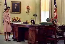 Johnson sitting behind the desk on the phone while Lady Bird Johnson stands to the side dressed in pink.