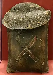 A brown leather case with a large oval flap and a diagonal cross, also in leather, stitched to the front