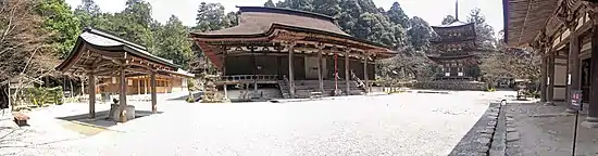 Color photograph of the enclosure of a Buddhist temple with, in the center, a brown wooden hall on one floor, at the back, a three-storey pagoda, on the left, an ablution pavilion and, in the foreground, a Light gray ground. Background: trees with green foliage.