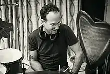 black-and-white image of White male wearing a dark t-shirt, glasses, and a set of headphones around his neck, sitting behind a drum kit, looking left of camera and laughing