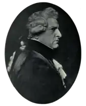 Right profile of stern-faced man in dark clothing with lacy shirt and cuffs, wearing a wig