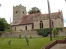 A stone church with red tiled roofs seen from the south. Extending from the chancel on the right is a battlemented chapel and, to the left, the tower is also battlemented