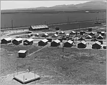 A black and white image shows approximately 20 tents or trailers lined up in a field next to pea field that is being harvested. The largest tent is in the very back and is an emergency community school for migrant children. The pea fields and some mountains take up the rest of the background.