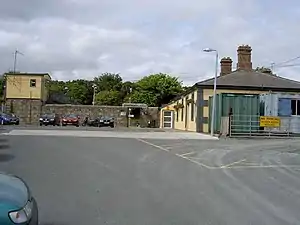 Photo of entrance of Wicklow Train Station taken from outside. The picture is taken during the day. The sky is blue, with white clouds. There are cars parked outside the station entrance. The station office (on the right hand side) is a yellow building (one storey) with green borders. There are trees on the background. The foreground is the road where the photo is taken from