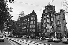 Occupied brick buildings with people standing outside, photographed from across the street